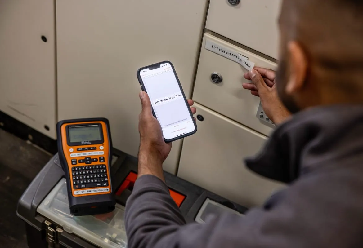 Electrician with a phone in hand prints a label on a Brother label printer beside a cabinet