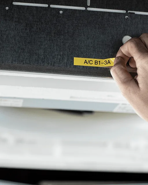 A hand fastens a yellow Brother label onto a cover in the ceiling